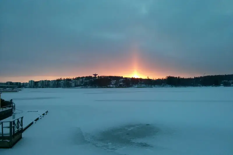 Aurinko nousee jäätyneen järven takaa, ja taivaalla on valopatsas.