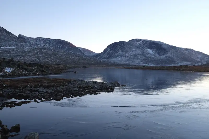 Ohut jää tyynen järven rantavedessä, taustalla lumisia tuntureita.