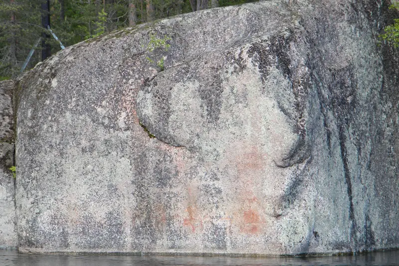 Pystysuora graniittikallio veden rajassa, pinnassa haaleita punaisia kuvioita.