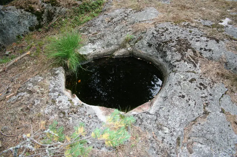 Pyöreä, tumman veden täyttämä kuoppa kalliossa. Reunoilla kasvaa heinätupsu ja männyntaimia.