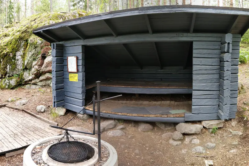 Harmaa hirsilaavu kallion vieressä, aukon edessä pyöreä tulisija ritilöineen.
