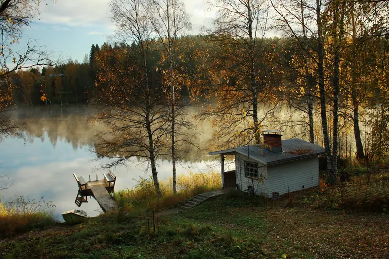 Syksyinen järvenranta aamulla: usva leijuu tyynen veden pinnalla, rannalla koivuja ja pieni sauna.