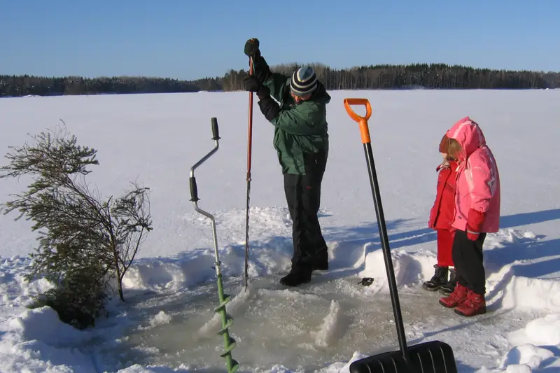 Aikuinen sahaa jäähän avantoa jääsahalla, lapsi katsoo vieressä; jääkaira ja lumilapio pystyssä.