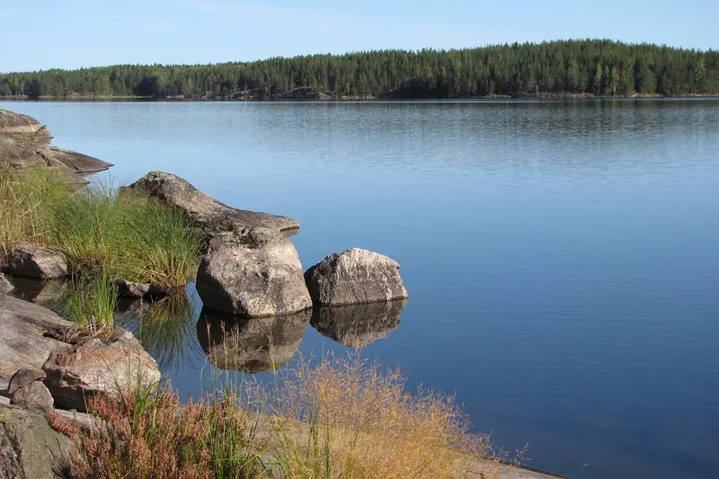 Isoja harmaita kiviä ja kalliota rannassa, kirkas tyyni vesi ja metsäinen vastaranta.