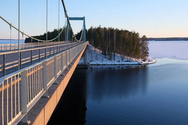 Riippusilta salmen yli. Sillan alla on avovettä, ja rannoilla on lunta ja jäätä.