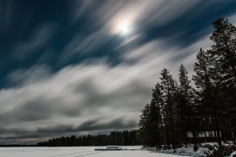 Kuu ohuen pilviharson takana talviyönä; alla luminen järvenjää, laituri ja rannan männyt mustina.